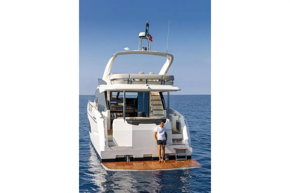 A man stands on the platform of a luxury yacht, anchored in calm blue waters under a clear sky.
